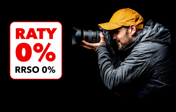 Mężczyzna w żółtej czapce i szarej kurtce trzyma aparat fotograficzny z dużym obiektywem, celując nim przed siebie. Obok znajduje się biały prostokąt z czerwonym napisem RATY 0% oraz czarnym tekstem RRSO 0%.